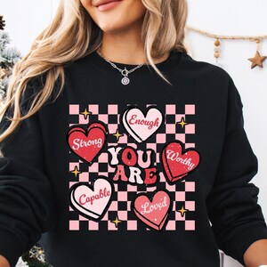 May include: Black sweatshirt with a pink and white checkered pattern and hearts that say "You Are" with words like "Strong", "Capable", "Enough", "Worthy", and "Loved" inside each heart.