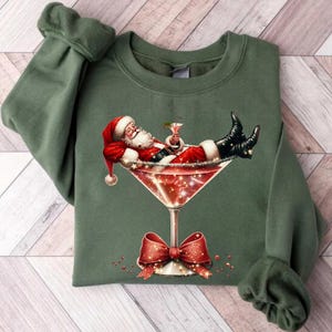 May include: A green sweatshirt with a festive design featuring Santa Claus relaxing in a martini glass with a red bow.