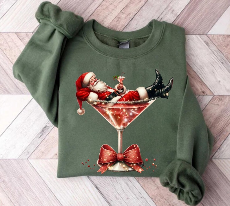 May include: A green sweatshirt with a festive design featuring Santa Claus relaxing in a martini glass with a red bow.