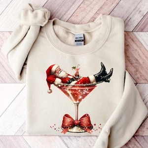 May include: A cream-colored sweatshirt with a festive design featuring Santa Claus relaxing in a martini glass with a red bow. The design is printed on the front of the sweatshirt.