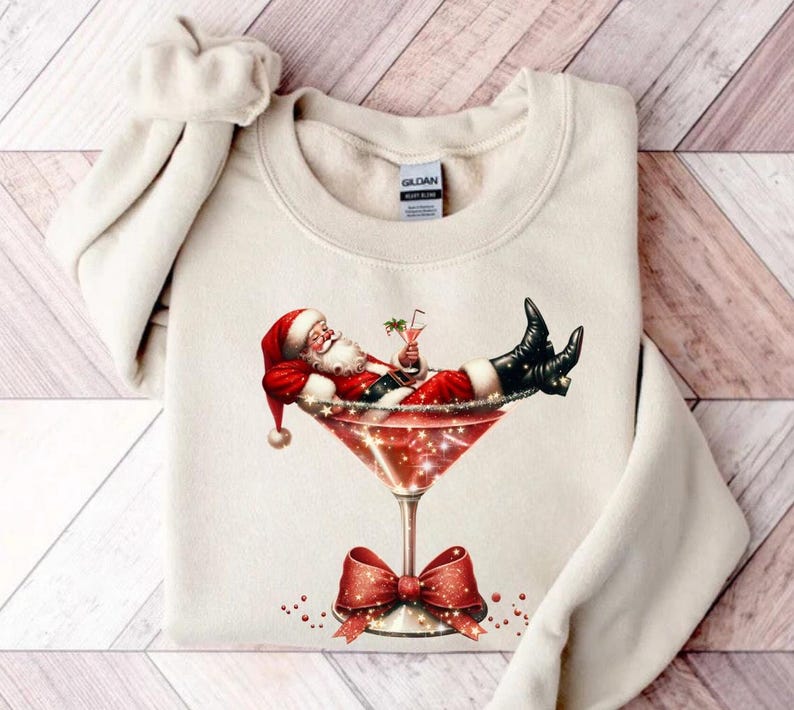 May include: A cream-colored sweatshirt with a festive design featuring Santa Claus relaxing in a martini glass with a red bow. The design is printed on the front of the sweatshirt.