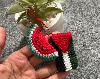 Könnte beinhalten: Ein gehäkelter Wassermelonen-Schlüsselanhänger mit einem palästinensischen Flaggen-Design. Der Schlüsselanhänger ist an einem silbernen Schlüsselring und einer roten Kette befestigt. Der Text "Für jeden Verkauf spenden wir an Palästina" ist im Bild sichtbar.