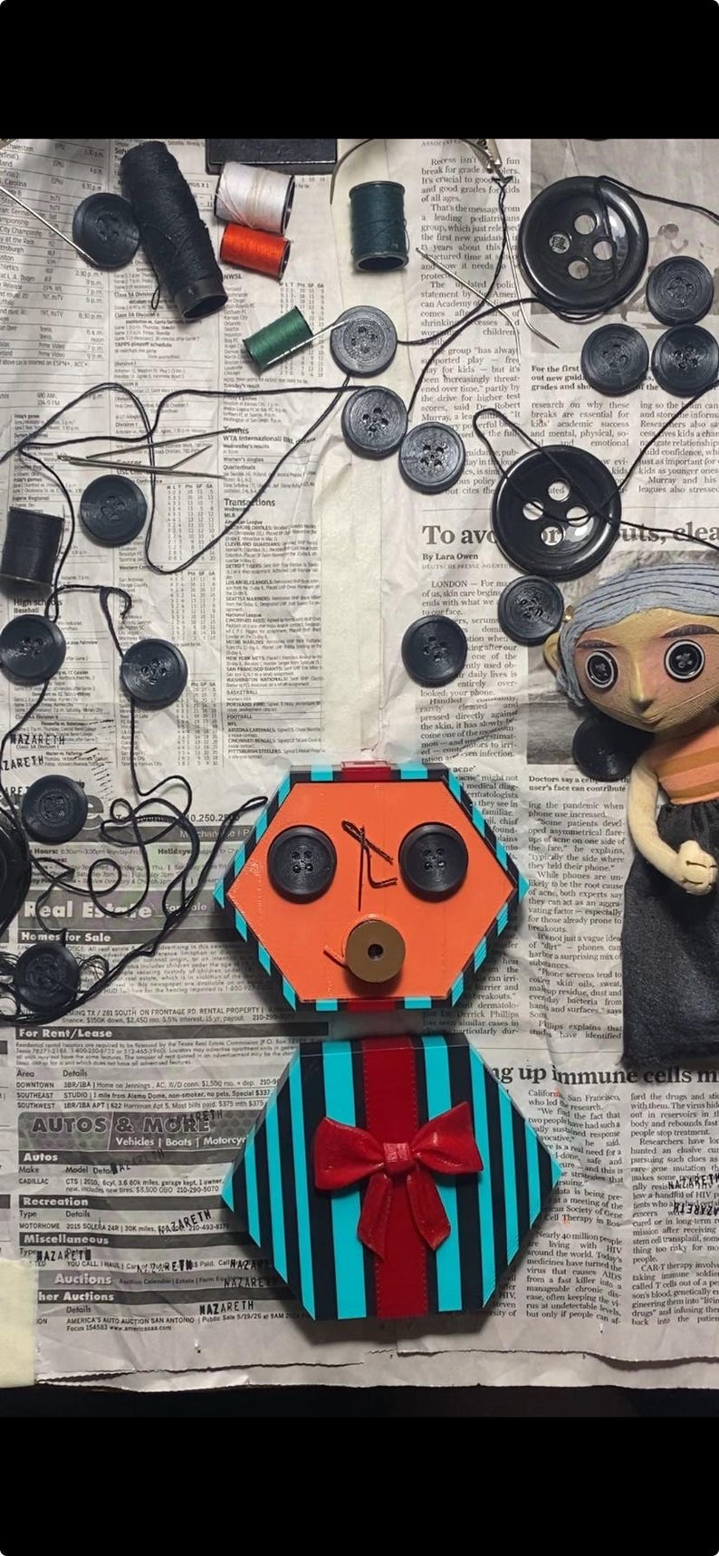 May include: A handmade doll with a hexagonal orange face featuring two black button eyes and a brown button nose, wearing a teal and black striped shirt with a red bow, accompanied by scattered black buttons, thread spools, and needles.