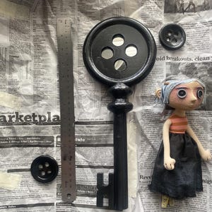 May include: A black key with a button-shaped head and a doll with button eyes. The key is next to a metal ruler and two black buttons. The doll has a striped shirt and a black skirt. The items are on a newspaper background.