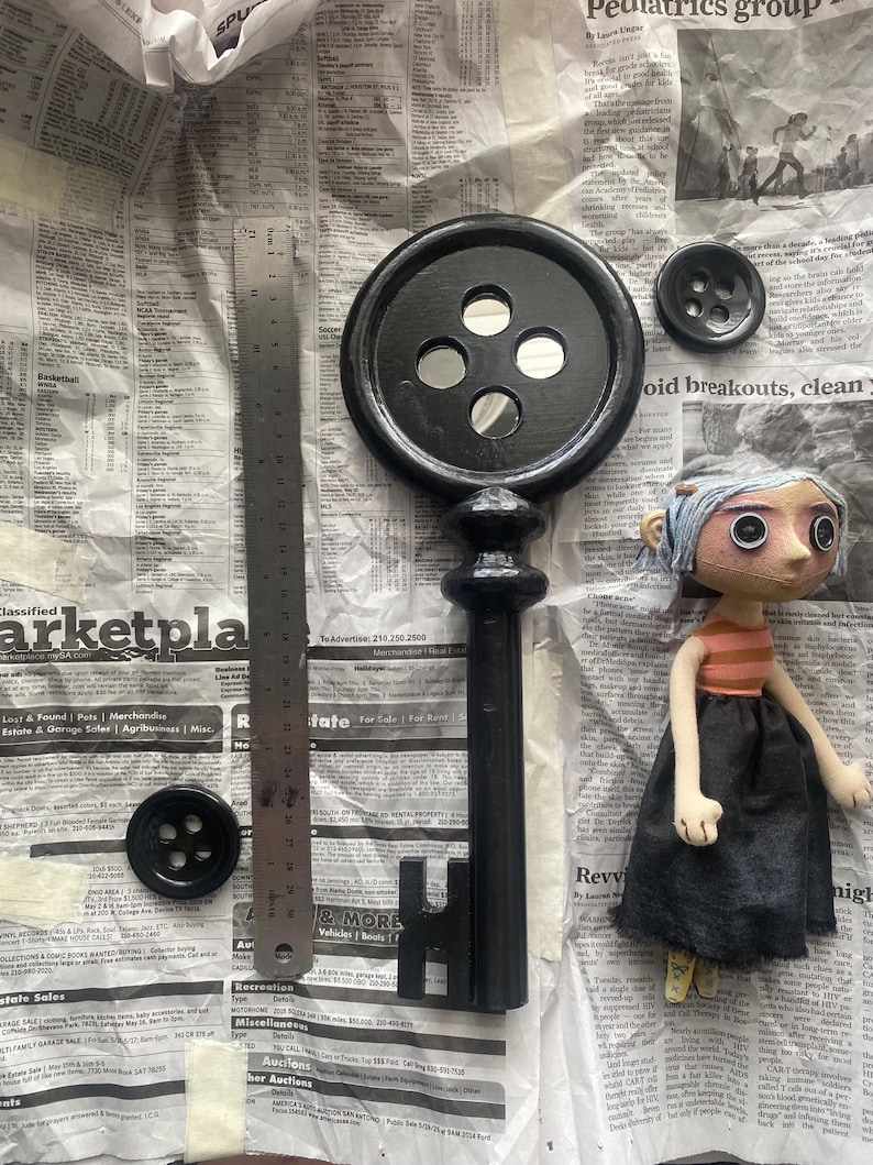 May include: A black key with a button-shaped head and a doll with button eyes. The key is next to a metal ruler and two black buttons. The doll has a striped shirt and a black skirt. The items are on a newspaper background.