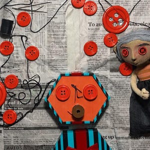 May include: A handmade doll with button eyes, a hexagonal orange face with button features, and a striped blue and black body with a red bow. The doll is surrounded by scattered red buttons, black thread, and spools of thread on a newspaper background.