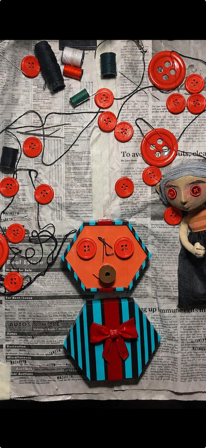 May include: A handmade doll with button eyes, a hexagonal orange face with button features, and a striped blue and black body with a red bow. The doll is surrounded by scattered red buttons, black thread, and spools of thread on a newspaper background.