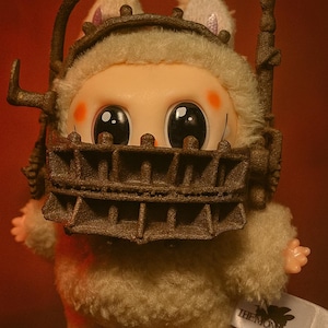 Puede incluir: Una figura de peluche con ojos grandes y oscuros y mejillas rosadas, que lleva una máscara y una diadema de metal marrón en forma de jaula. La figura está cubierta de suave pelaje color crema. La etiqueta dice "THE MONSIFRS".