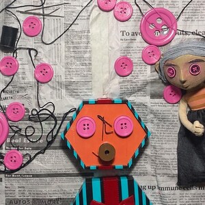 May include: A collection of pink buttons, sewing thread spools, and a doll with button eyes arranged on newspaper. Below, a hexagonal orange box with button eyes and a mouth, and a smaller box with blue and black stripes and a red bow.