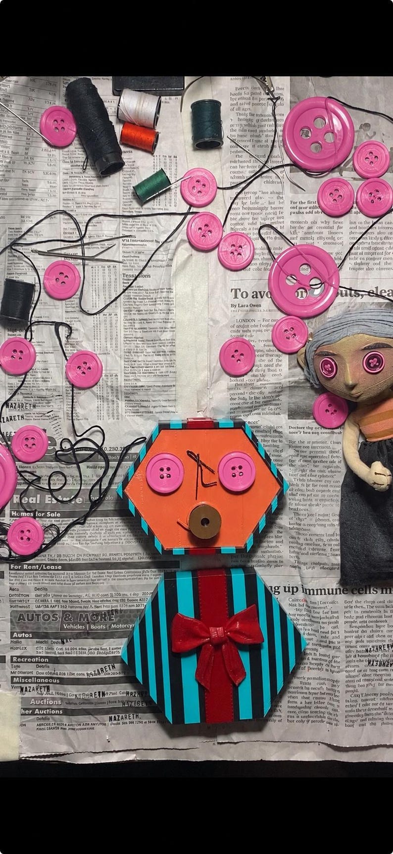 May include: A collection of pink buttons, sewing thread spools, and a doll with button eyes arranged on newspaper. Below, a hexagonal orange box with button eyes and a mouth, and a smaller box with blue and black stripes and a red bow.