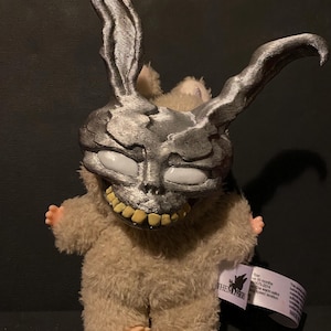 May include: A plush toy featuring a silver mask with a skeletal design and long rabbit ears. The toy has a tan, furry body, small hands and feet, and a tag that reads "THE MONSTERS". The mask has white eyes and yellow teeth.