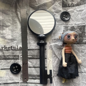 May include: A black key-shaped mirror with a round frame, a doll with a striped shirt and black skirt, and two black buttons are displayed on a newspaper background. A ruler is also present. The items are likely part of a craft or art project.
