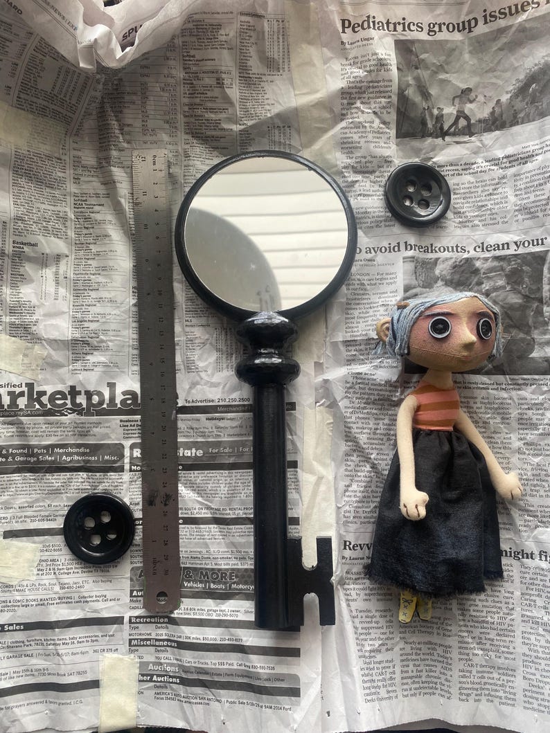 May include: A black key-shaped mirror with a round frame, a doll with a striped shirt and black skirt, and two black buttons are displayed on a newspaper background. A ruler is also present. The items are likely part of a craft or art project.