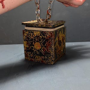 May include: A black and gold decorative box with a zipper closure and a chain handle. The box has a geometric pattern and is embellished with gold glitter. The box is connected to a chain that is attached to a wrist.