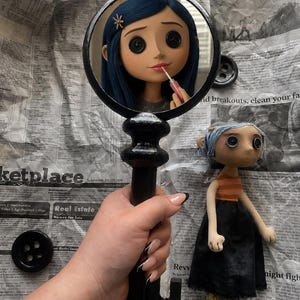 May include: A black key-shaped mirror reflects a doll with blue hair and button eyes applying lipstick. A doll with blue hair, an orange striped shirt, and a black skirt stands nearby. The items are on a newspaper background.