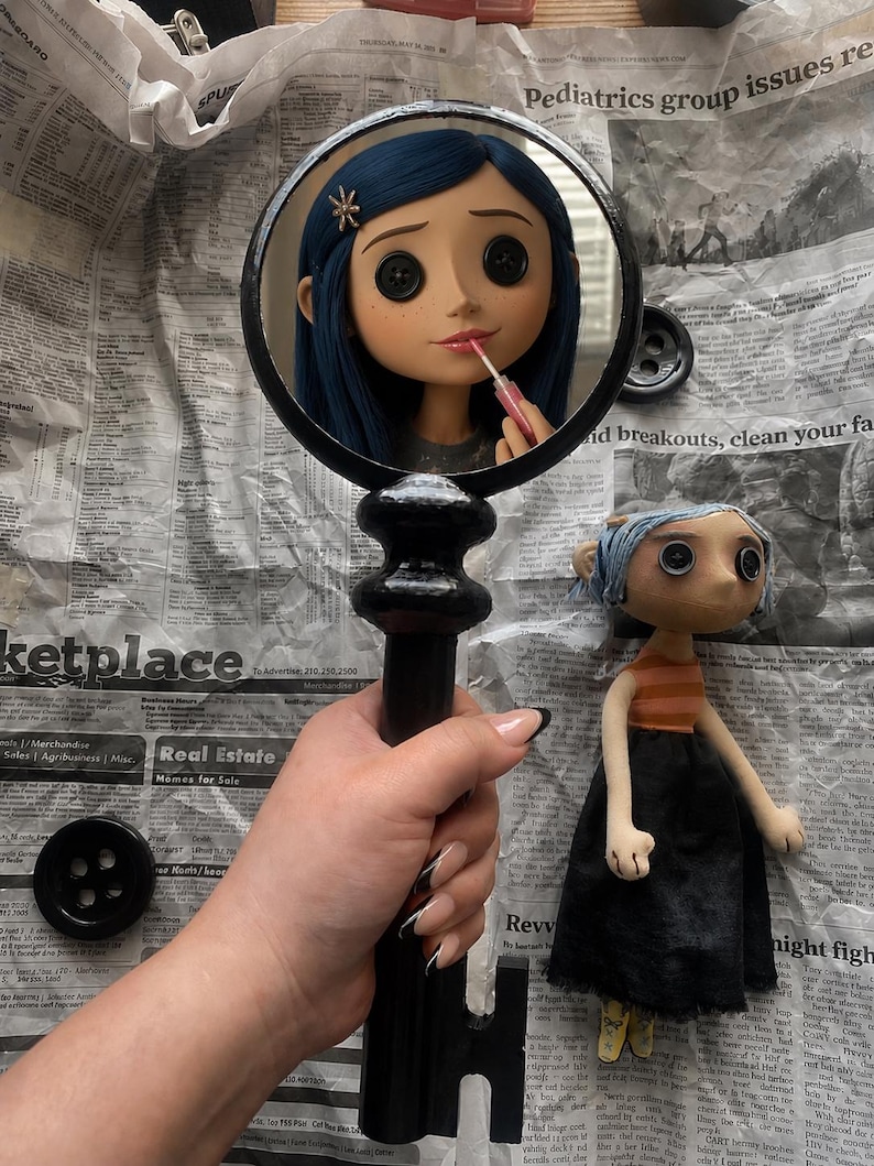 May include: A black key-shaped mirror reflects a doll with blue hair and button eyes applying lipstick. A doll with blue hair, an orange striped shirt, and a black skirt stands nearby. The items are on a newspaper background.