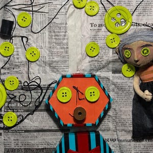 May include: A handmade doll with button eyes and a hexagonal orange face featuring two green buttons as eyes and a brown button as a mouth. The doll has a blue and black striped body with a red bow and is surrounded by scattered green buttons and thread.