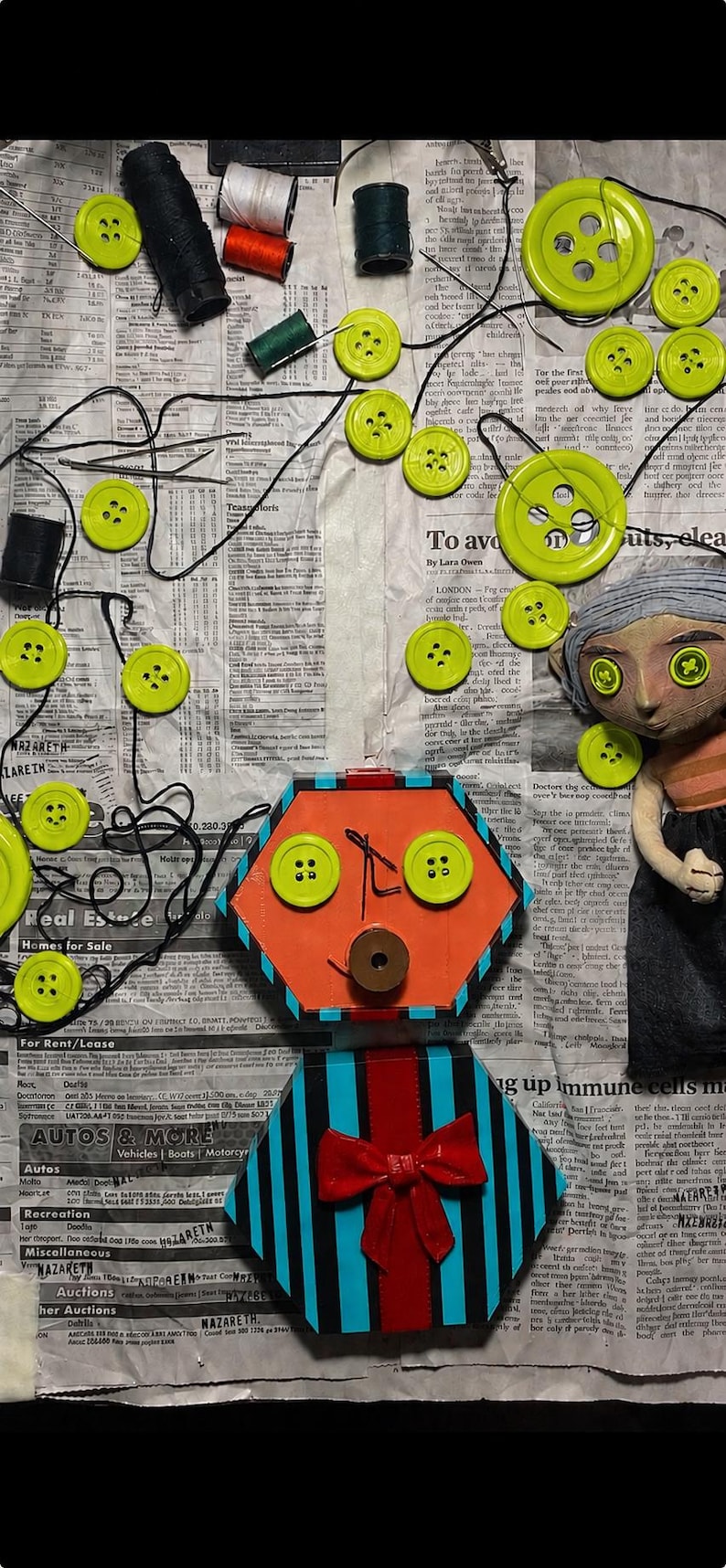 May include: A handmade doll with button eyes and a hexagonal orange face featuring two green buttons as eyes and a brown button as a mouth. The doll has a blue and black striped body with a red bow and is surrounded by scattered green buttons and thread.