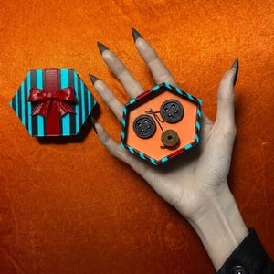 May include: A hexagonal gift box with a teal and black striped pattern and a red bow on the lid. Inside the open box are two black four-hole buttons and one small brown spool of thread. The box features a bright orange interior base.