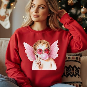 May include: Red sweatshirt with a graphic of a cherub wearing pink sunglasses and blowing a bubble. The cherub has pink wings and is holding a peace sign.