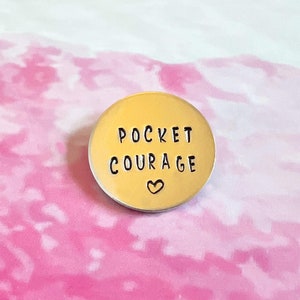 May include: A gold-colored metal disc with the words "Pocket Courage" and a small heart symbol engraved on it.