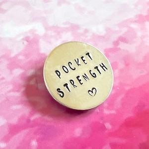 以下が含まれることがあります： 「ポケットストレングス」という言葉と小さなハートのシンボルが刻まれた銀色の丸いチャーム