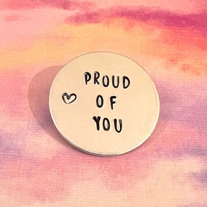 May include: A circular silver-coloured metal token featuring the stamped black text PROUD OF YOU arranged in three lines, accompanied by a small stamped heart symbol to the left of the word PROUD. The surface of the metal disc is smooth and reflective.