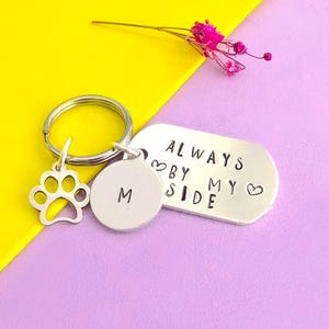 May include: A silver keychain with a paw print charm, a round charm with the letter "M", and a rectangular tag that reads "ALWAYS BY MY SIDE" with heart symbols. The keychain is on a yellow and purple background.