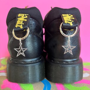 May include: Black platform boots with a silver star charm hanging from a metal ring. The boots feature a yellow and black tag that says "Air Wair". The background is pink.