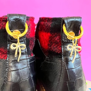 May include: Close-up of two black ankle boots with red and black plaid fabric on the back. The boots have gold-coloured accents, including a bow-shaped charm. The boots have a faux crocodile skin texture.