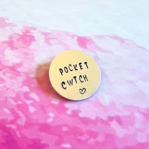May include: A circular gold-toned metal disc featuring the stamped black text POCKET CWTCH and a small heart symbol centred below the text. The metal surface has a smooth, reflective finish with clearly legible, dark, uppercase lettering.