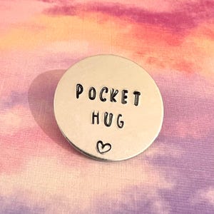 May include: A circular silver-coloured metal token featuring the stamped black text POCKET HUG and a small heart symbol centred below the words. The surface of the metal disc is smooth and reflective with a clean, minimalist design.