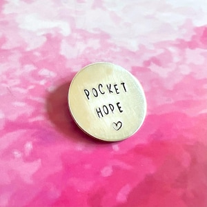 May include: A circular silver-coloured metal token featuring the stamped black text POCKET HOPE and a small heart symbol centred below the words. The token has a smooth, brushed metallic finish and a clean, simple design on its front surface.