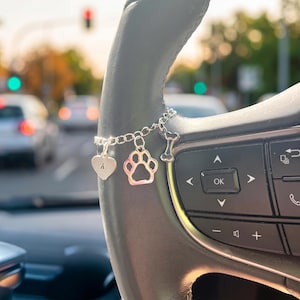 May include: A silver-toned metal charm bracelet featuring three distinct charms: a heart-shaped pendant engraved with the letter A, a cutout paw print, and a small dog bone. The bracelet is draped over the steering wheel of a vehicle.