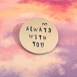 May include: A circular silver-toned metal token featuring the stamped black text "ALWAYS WITH YOU" arranged in three lines, with a small heart symbol positioned above the word "ALWAYS" on the smooth, reflective surface of the disc.