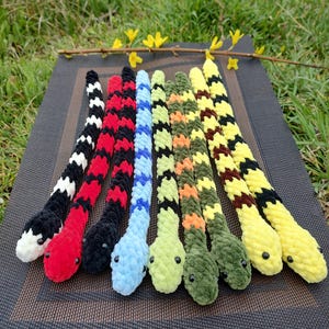 May include: A set of nine crocheted snake toys in various colours, including black and white, red and black, blue and white, green and yellow, and yellow. The snakes are arranged in a row on a black and grey textured surface.