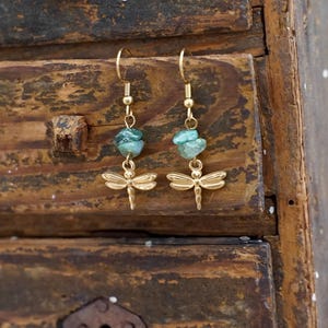 Pode incluir: Um par de brincos dourados com libélulas e contas de pedra de cor verde-azulado. Os brincos estão pendurados numa superfície de madeira. Os brincos têm um fecho de gancho.