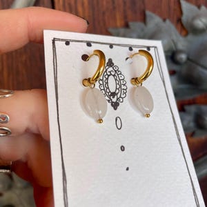May include: A pair of gold hoop earrings with white stone drops. The earrings are displayed on a white card with black polka dots.
