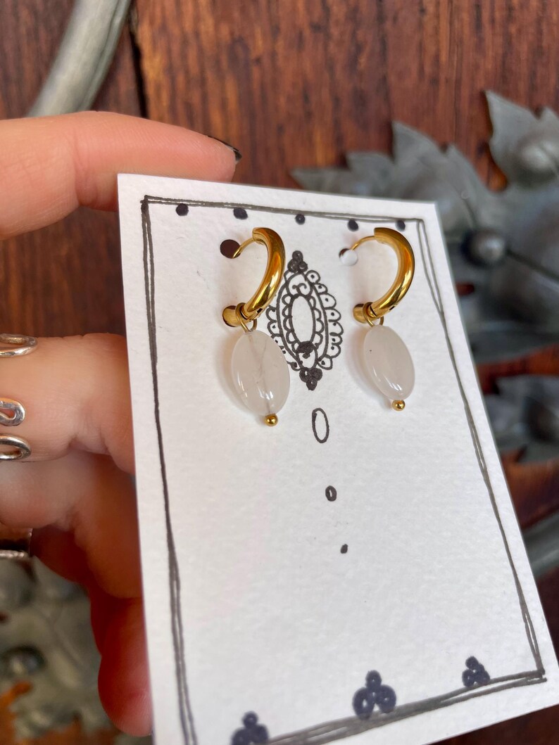 May include: A pair of gold hoop earrings with white stone drops. The earrings are displayed on a white card with black polka dots.