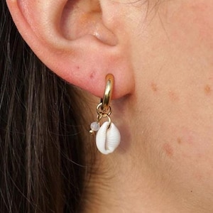 May include: A gold hoop earring featuring a small white seashell pendant and a small, light-coloured bead. The earring is worn on an ear, with the focus on the jewellery.