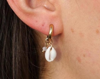 May include: A gold hoop earring with a small white seashell pendant and a small, light-colored bead. The earring is worn on a person's ear, with the focus on the jewelry.