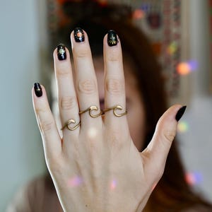 May include: A hand wearing black nail polish with gold accents and three gold wave rings.