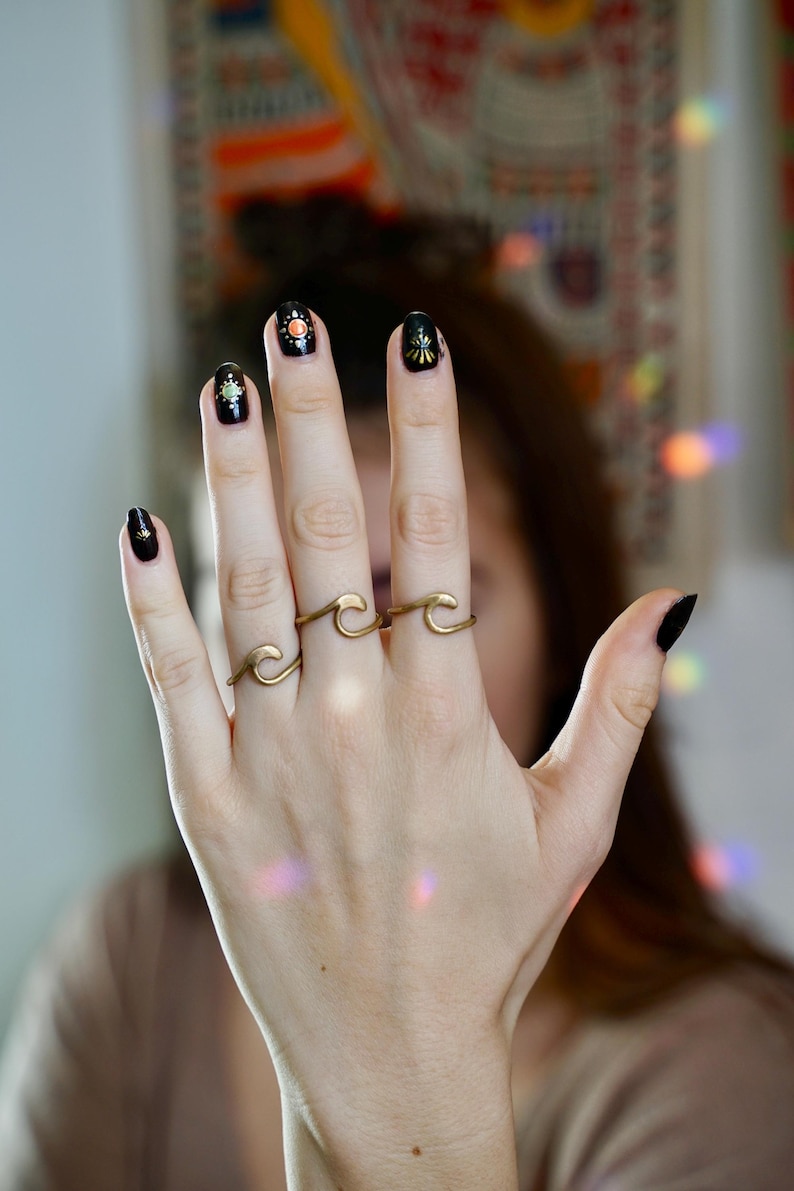 May include: A hand wearing black nail polish with gold accents and three gold wave rings.