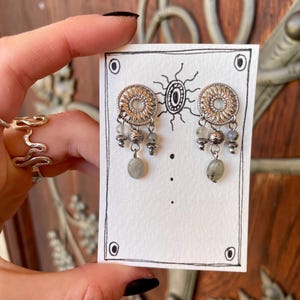 Pode incluir: Brincos prateados com um design circular vazado e contas cinzentas penduradas. Os brincos s&atilde;o exibidos em um cart&atilde;o branco com uma ilustra&ccedil;&atilde;o de sol desenhada &agrave; m&atilde;o e pontos pretos. O fundo &eacute; uma superf&iacute;cie de madeira.