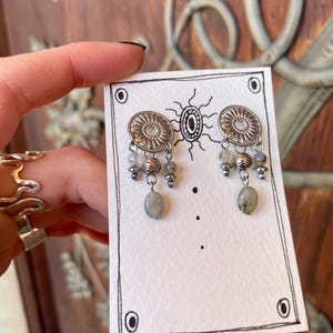 Pode incluir: Brincos prateados com um design circular e contas cinzentas penduradas. Os brincos s&atilde;o exibidos em um cart&atilde;o branco com uma ilustra&ccedil;&atilde;o preta e uma borda decorativa. O fundo &eacute; uma porta de madeira.