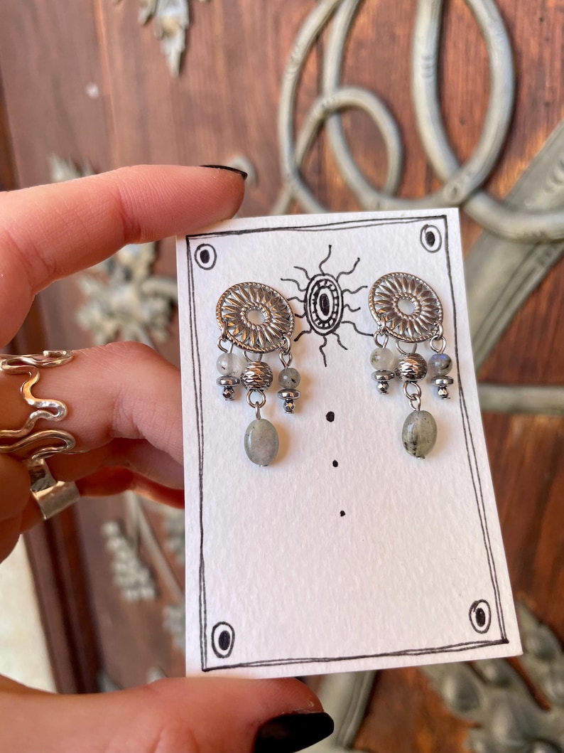 Pode incluir: Brincos prateados com um design circular e contas cinzentas penduradas. Os brincos s&atilde;o exibidos em um cart&atilde;o branco com uma ilustra&ccedil;&atilde;o preta e uma borda decorativa. O fundo &eacute; uma porta de madeira.