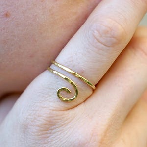Pode incluir: Um anel de cor dourada com uma banda de metal fina e martelada que envolve o dedo num design em espiral. O anel tem um acabamento polido e uma estrutura delicada e aberta que se curva numa pequena argola circular na parte frontal.