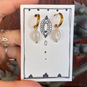 May include: A pair of gold hoop earrings with white oval-shaped beads hanging from them. The earrings are displayed on a white card with black ink drawings.
