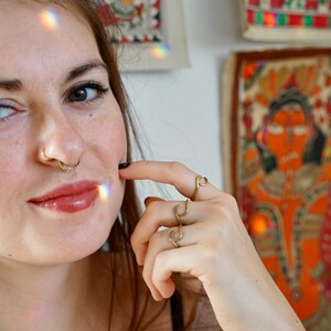 May include: A close-up of a person's face with a nose ring and a gold ring on their finger. The person is wearing a brown shirt and has long brown hair.