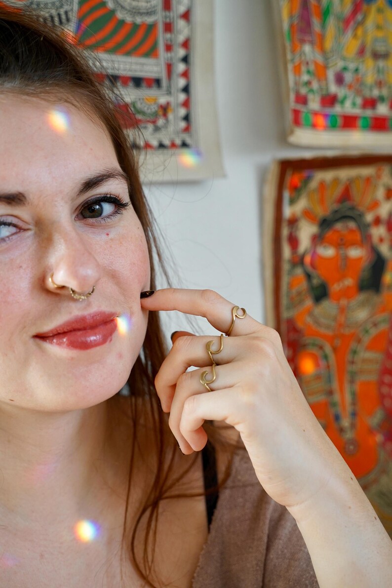 May include: A close-up of a person's face with a nose ring and a gold ring on their finger. The person is wearing a brown shirt and has long brown hair.